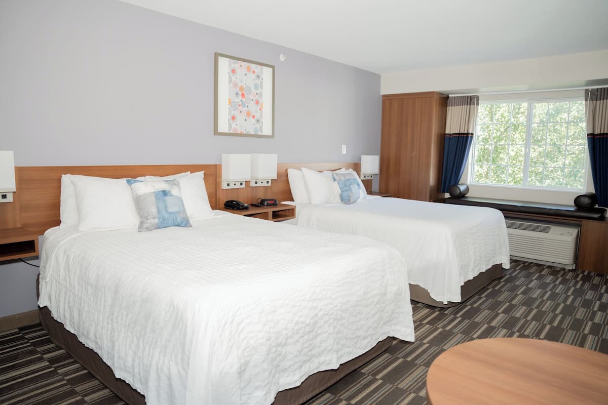 A hotel room with two beds, a small table, a window with curtains, and a wall-mounted artwork.