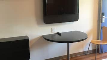 A hotel room with a flat-screen TV mounted on the wall, a small round table, a chair, a wooden dresser, and a bed with a red and white blanket.