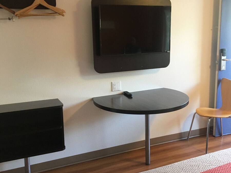 A hotel room with a flat-screen TV mounted on the wall, a small round table, a chair, a wooden dresser, and a bed with a red and white blanket.