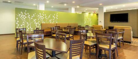 A hotel dining area with multiple tables and chairs, a green wall with a white tree design, and a flat-screen TV mounted on the wall.