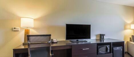 A hotel room with a dark wooden desk, a flat-screen TV, a microwave, a mini-fridge, a chair, and a lamp.