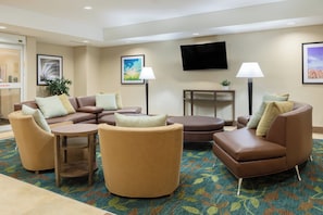 Un vestíbulo de hotel con un sofá modular, sillones, una mesa de centro, un televisor de pantalla plana y una pequeña mesa con una planta.