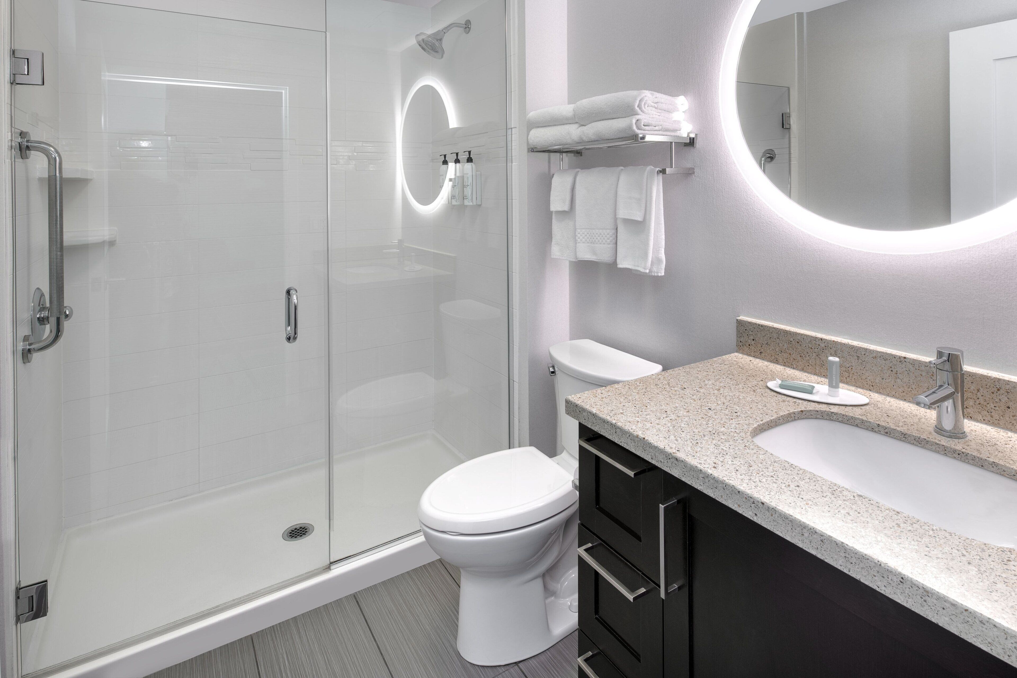 Un baño moderno con ducha acristalada, inodoro blanco, tocador de granito y un espejo grande.