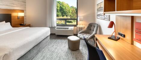 Et hotelværelse med en stor seng, et skrivebord, en stol, en lille skammel, en skrivebordslampe, et vindue med udsigt over grønne områder og en vægmonteret hylde.