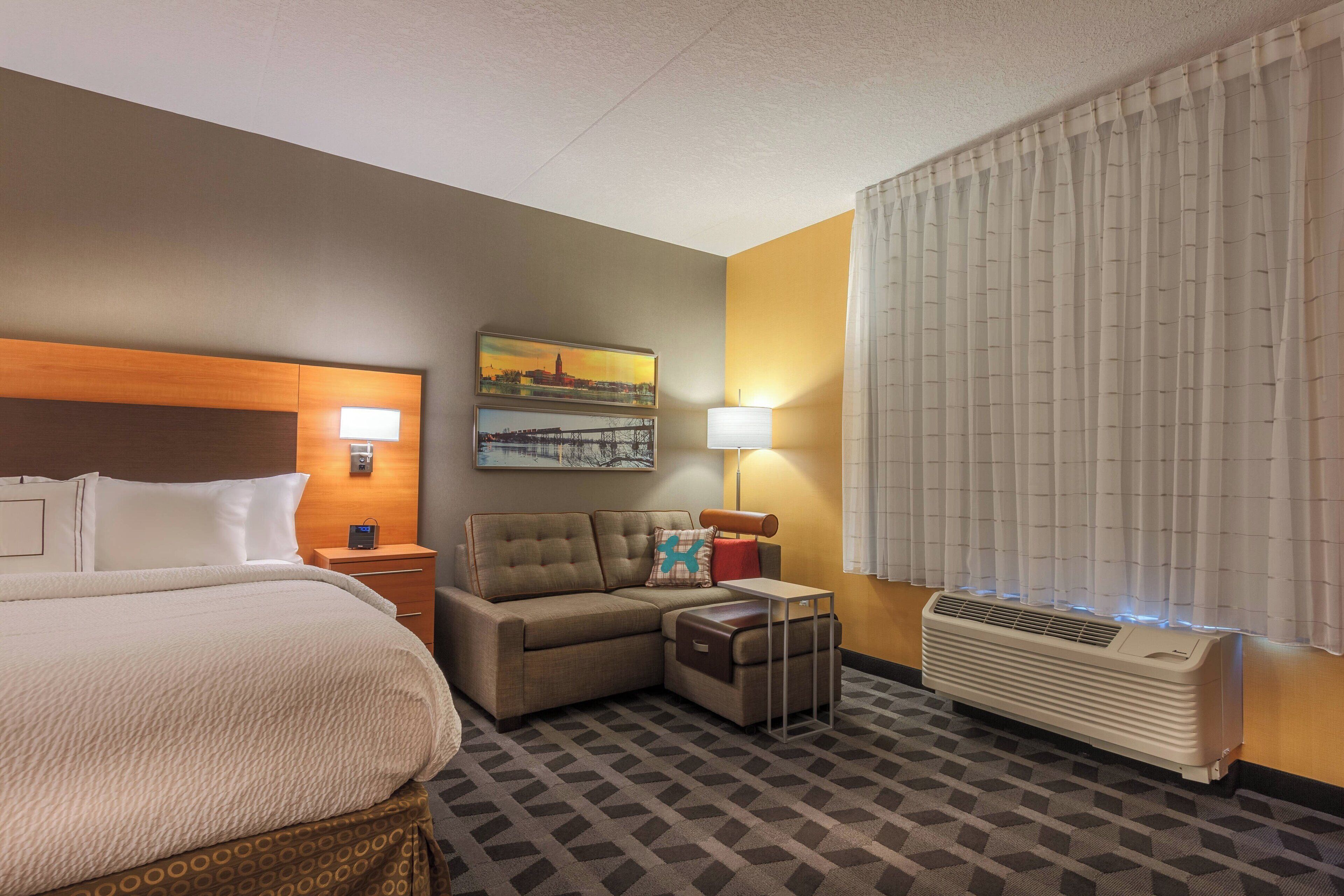 A hotel room with a bed, a sofa, a small table, a lamp, a heater, and a painting on the wall.