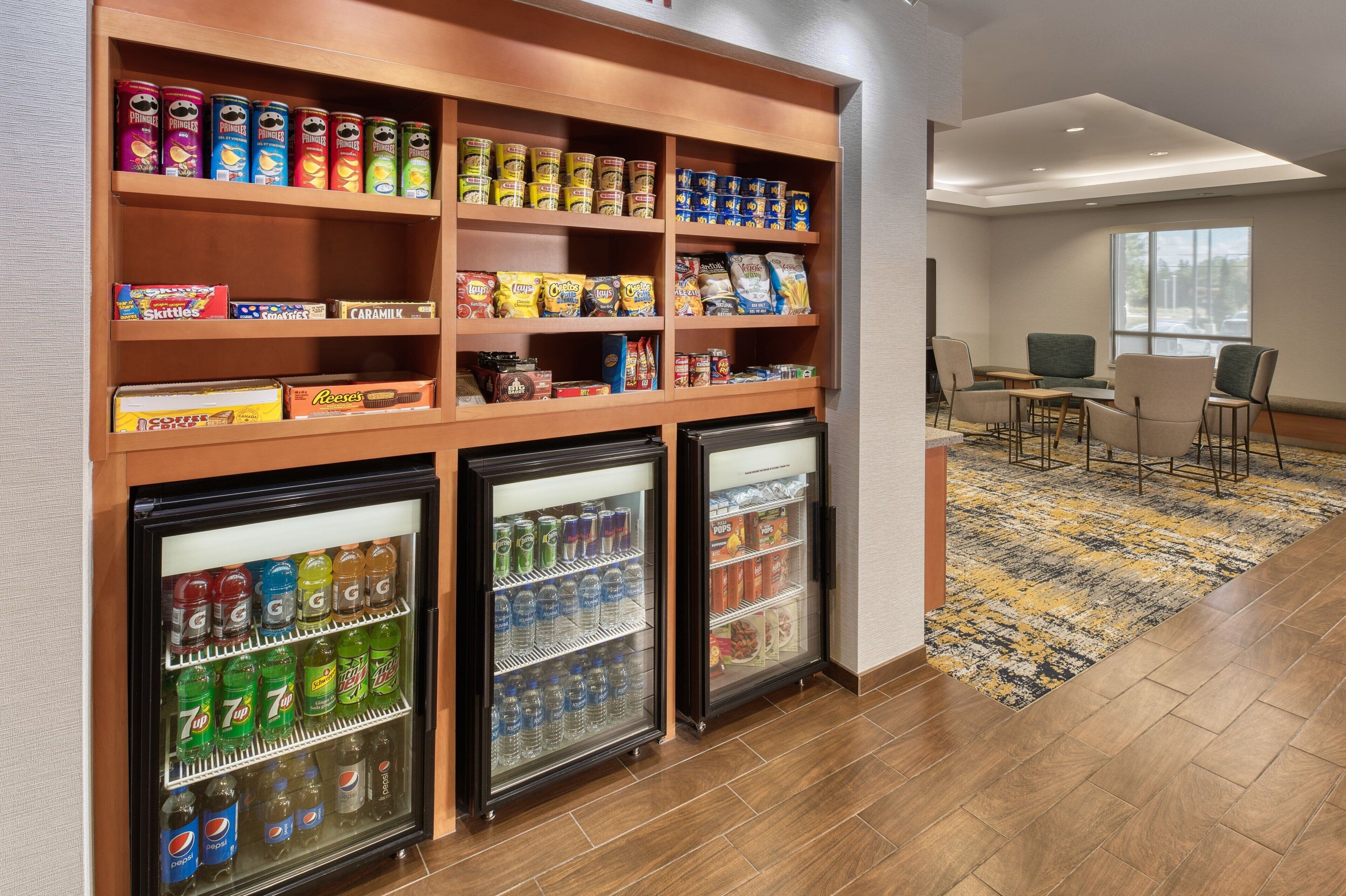 Um saguão de hotel com uma prateleira de lanches recheada de diversas opções e um refrigerador com bebidas.