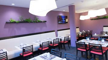 Ein Restaurant mit Tischen, Stühlen, einem Flachbildfernseher an der Wand und dekorativen Pflanzen auf Regalen.