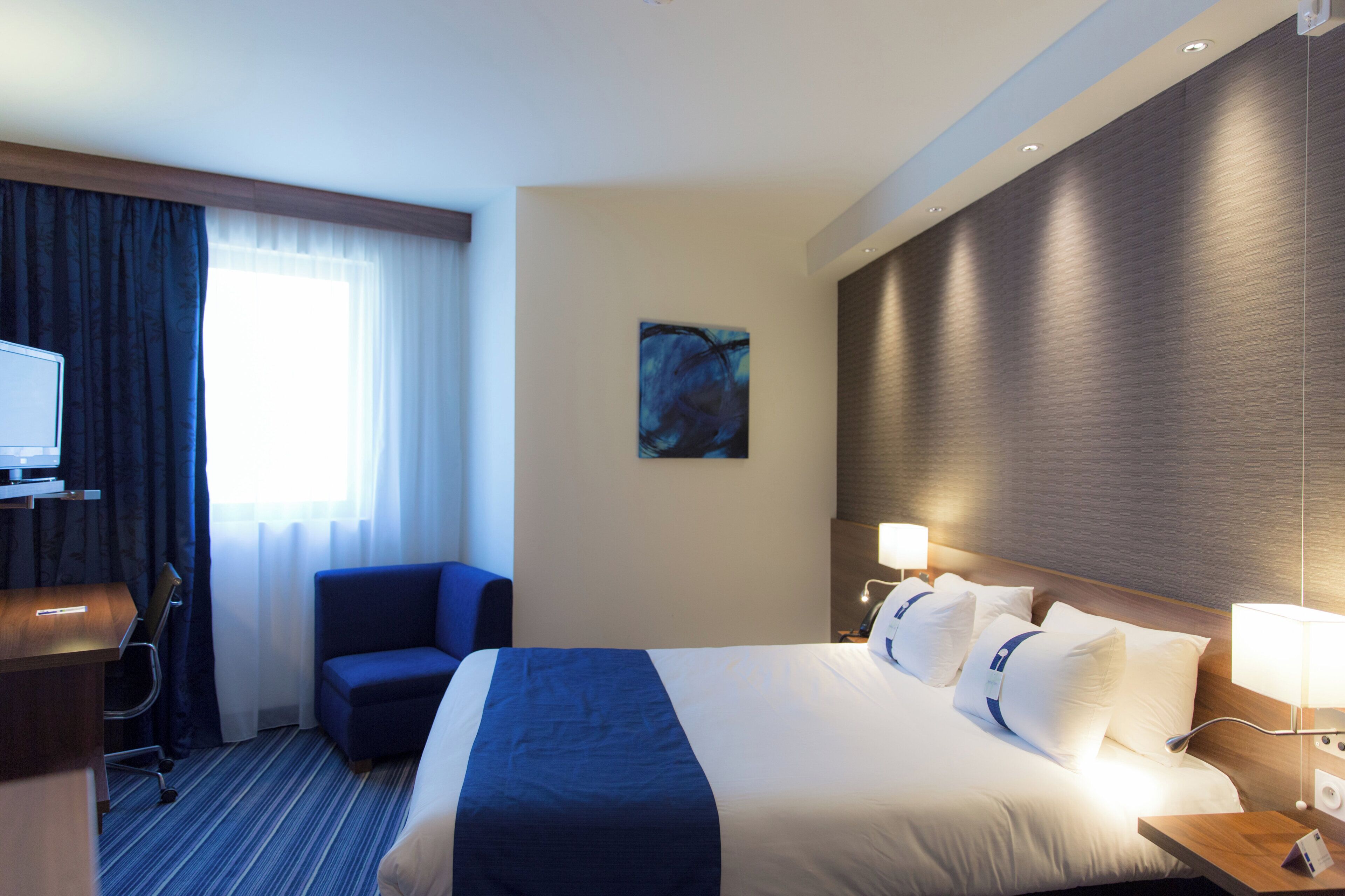 A hotel room with a bed, a desk, a chair, a television, and a painting on the wall.