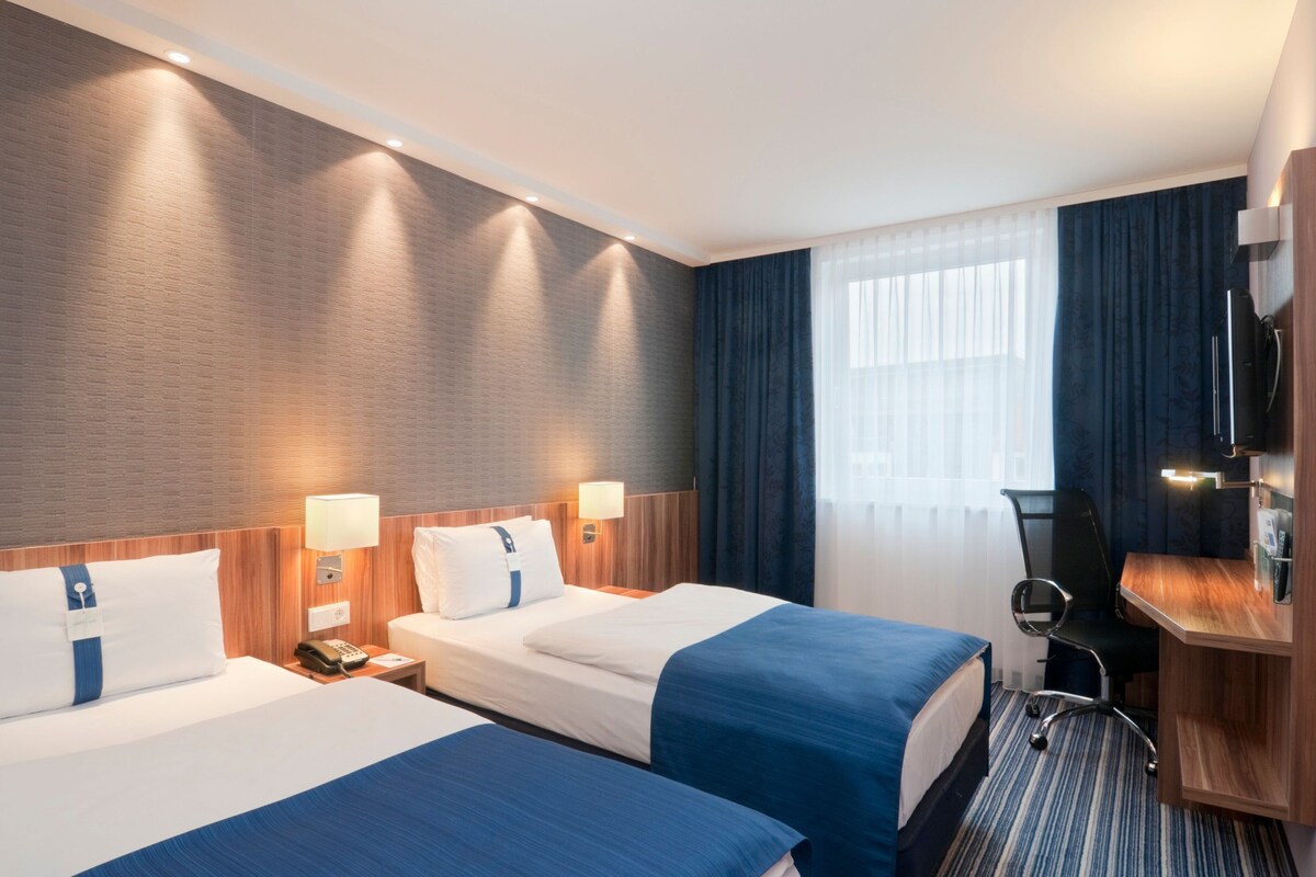 A hotel room with two beds, a desk, a TV, and a window with curtains.