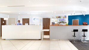 A modern hotel reception area with a white reception desk, a bar with various bottles, and two black bar stools. - Holiday Inn Express Toulon - Est by IHG (Toulon)
