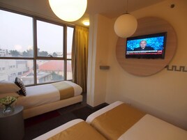 Habitación de hotel con dos camas, un televisor montado en la pared y vistas a la ciudad a través de amplios ventanales.