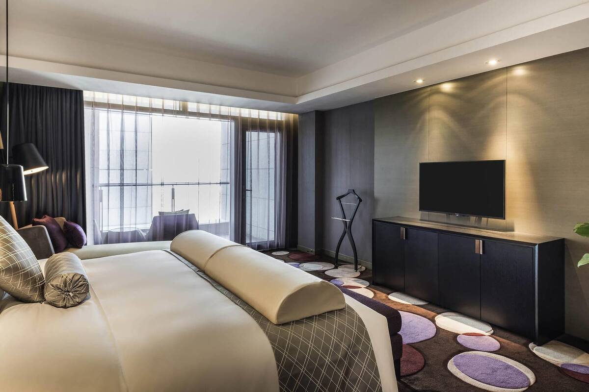 Una habitación de hotel moderna con una cama grande, un televisor de pantalla plana, un aparador de madera oscura y un ventanal con cortinas.