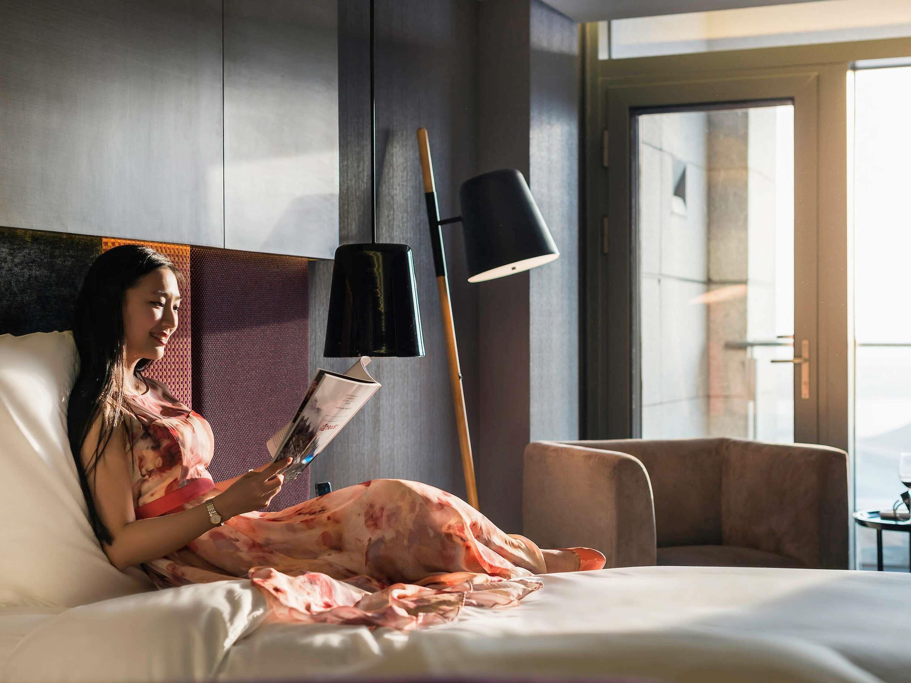 Una persona acostada en una cama en una habitación con un ventanal grande, una lámpara de pie y una silla.