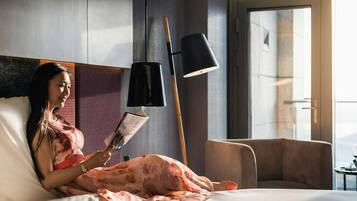 Una persona acostada en una cama en una habitación con un ventanal grande, una lámpara de pie y una silla.
