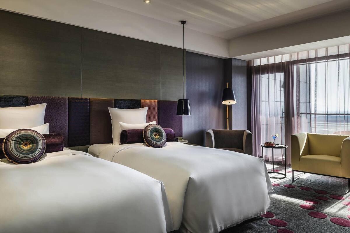 Habitación de hotel con dos camas, un área de descanso con una silla, una mesita y un ventanal amplio.