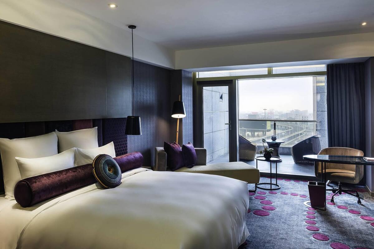 Habitación de hotel con una cama grande, un escritorio, una silla y vistas a la ciudad a través de la ventana.