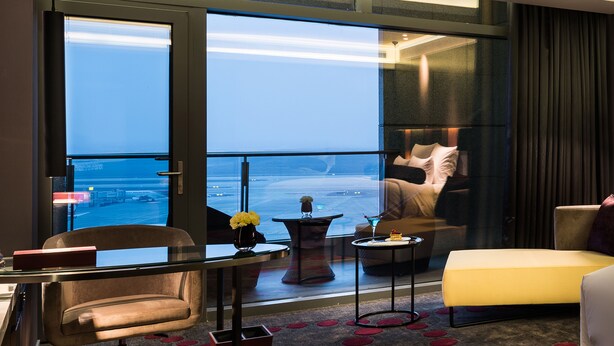 Habitación de hotel con un ventanal grande, una cama, un sillón amarillo, una mesita con un florero y vista al océano.