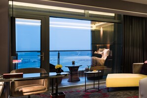 Habitación de hotel con un ventanal grande, una cama, un sillón amarillo, una mesita con un florero y vista al océano.
