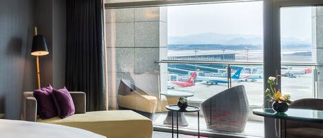 A hotel room with a large window, a bed, a chair, a small table, and a lamp.