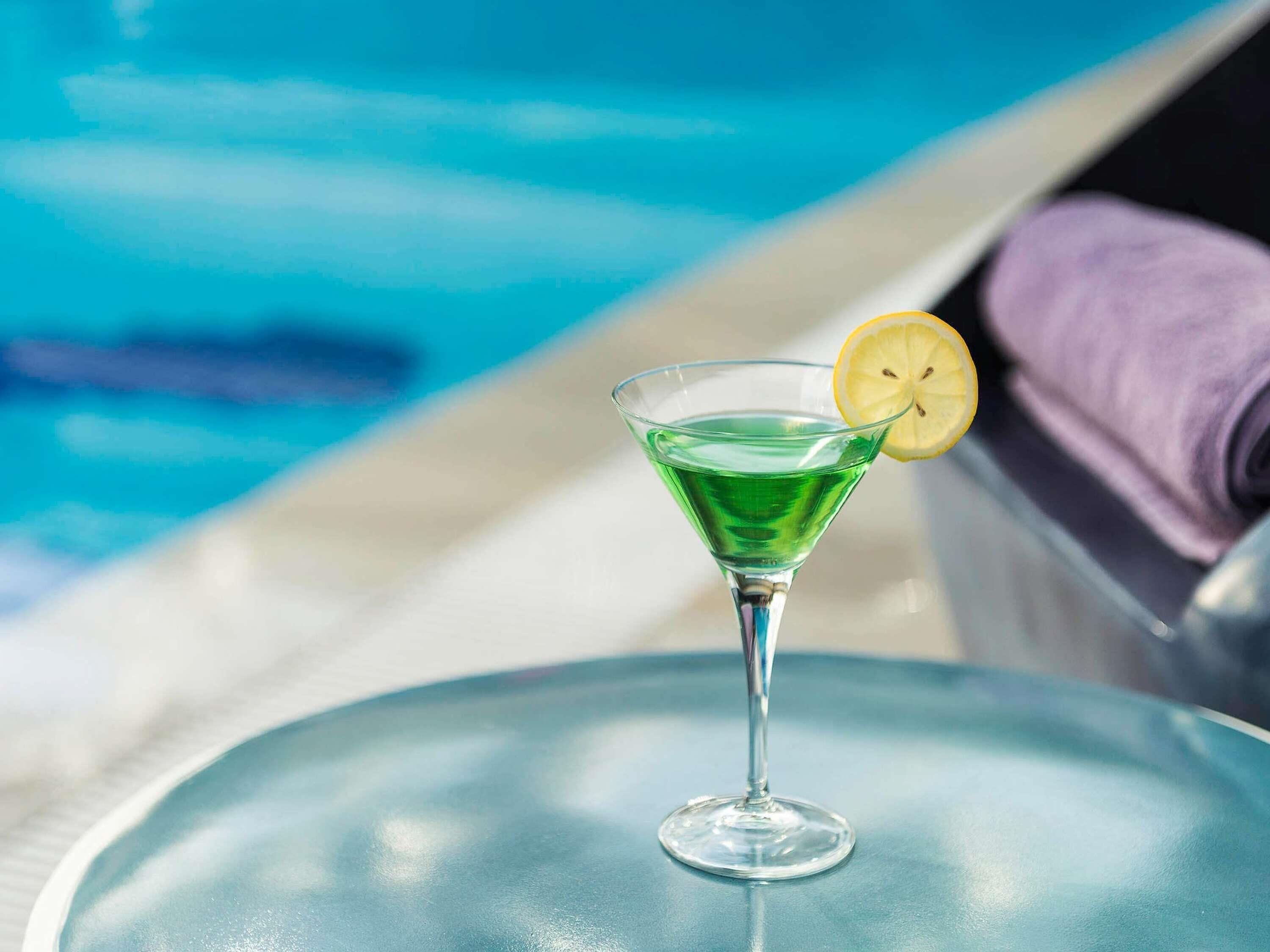 A clear glass martini glass with a lemon slice on a table by a poolside.