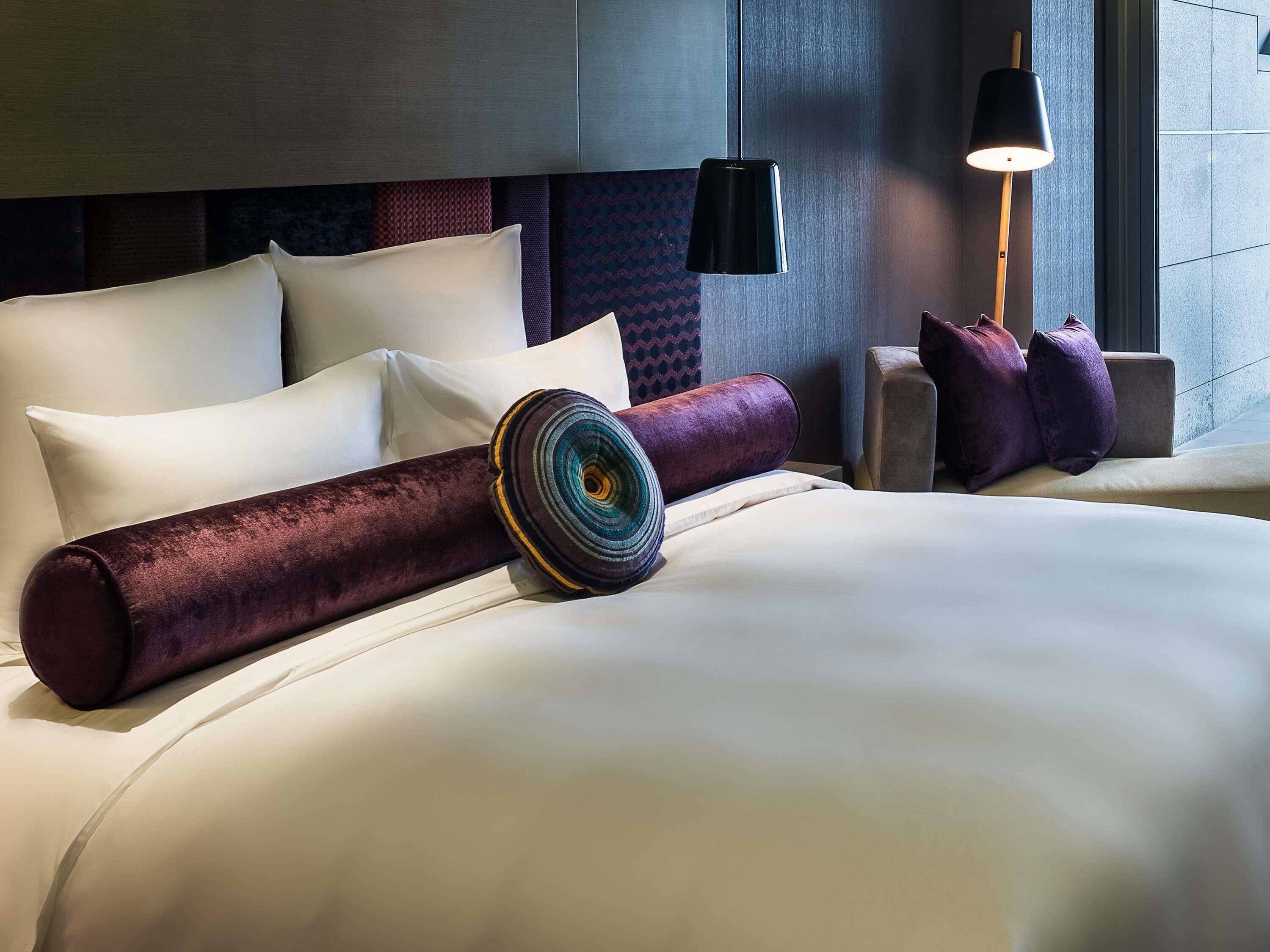 Una habitación de hotel moderna con una cama grande, un cabecero con diseño estampado, una mesita de noche con lámpara y un ventanal con vistas.
