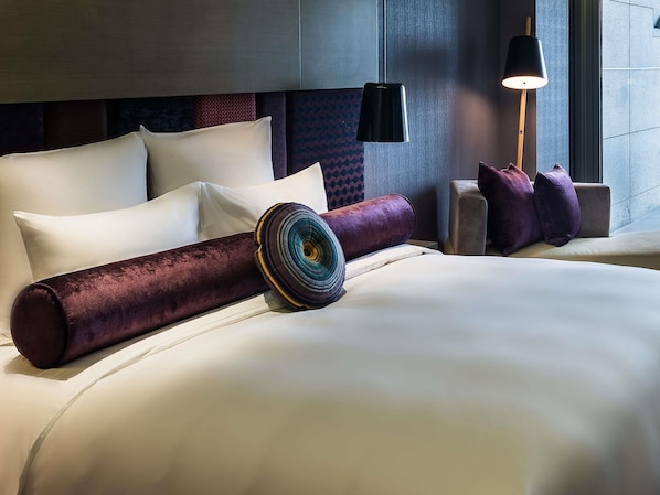 Una habitación de hotel moderna con una cama grande, un cabecero con diseño estampado, una mesita de noche con lámpara y un ventanal con vistas.