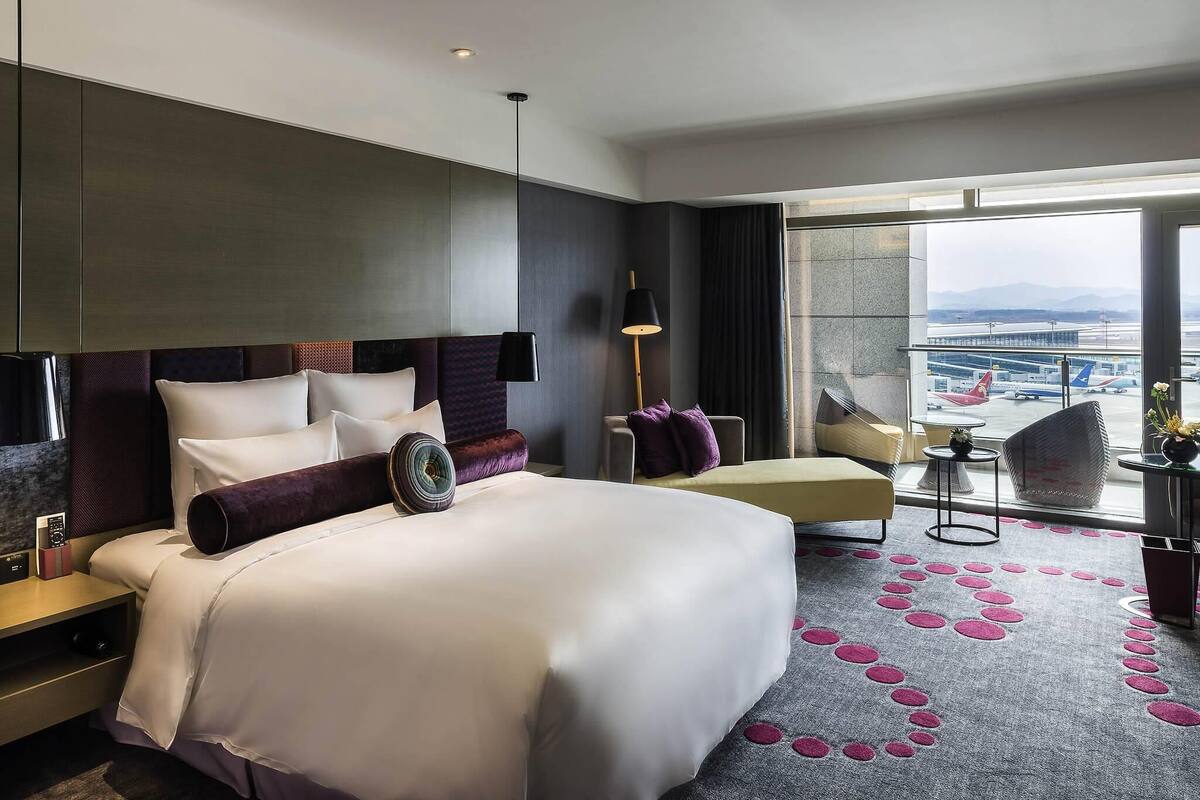 Una habitación de hotel moderna con una cama grande, una zona de estar con vistas al aeropuerto y un balcón también con vistas al aeropuerto.