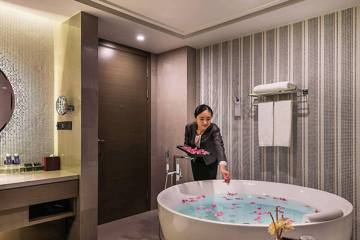 Un baño moderno con una bañera grande llena de agua y pétalos de rosa, un tocador con espejo y artículos de aseo, y un perchero con toallas blancas.