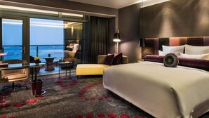 Una habitación de hotel moderna con una cama grande, un área de descanso con un sofá y sillones, una mesita y vistas al mar a través de amplios ventanales.