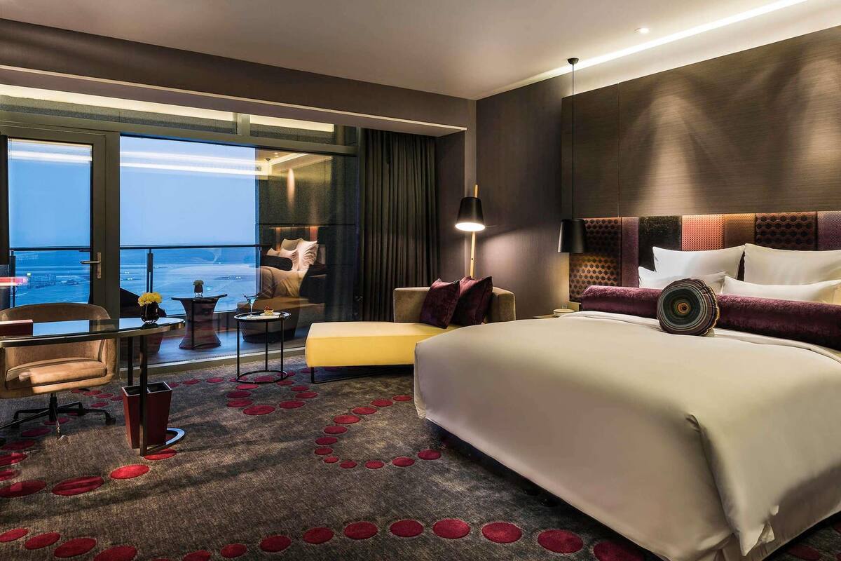Una habitación de hotel moderna con una cama grande, un área de descanso con un sofá y sillones, una mesita y vistas al mar a través de amplios ventanales.