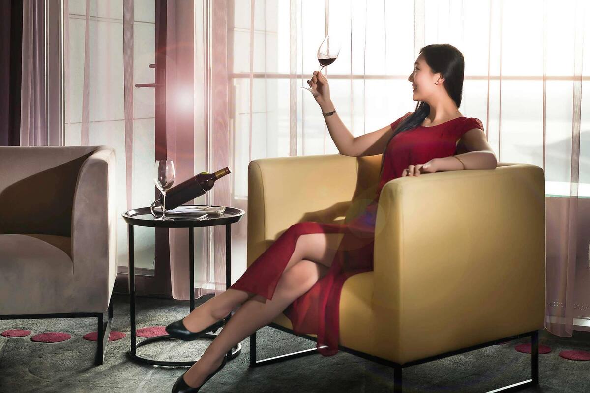 Una mujer con un vestido rojo sentada en un sillón amarillo, sosteniendo una copa de vino.