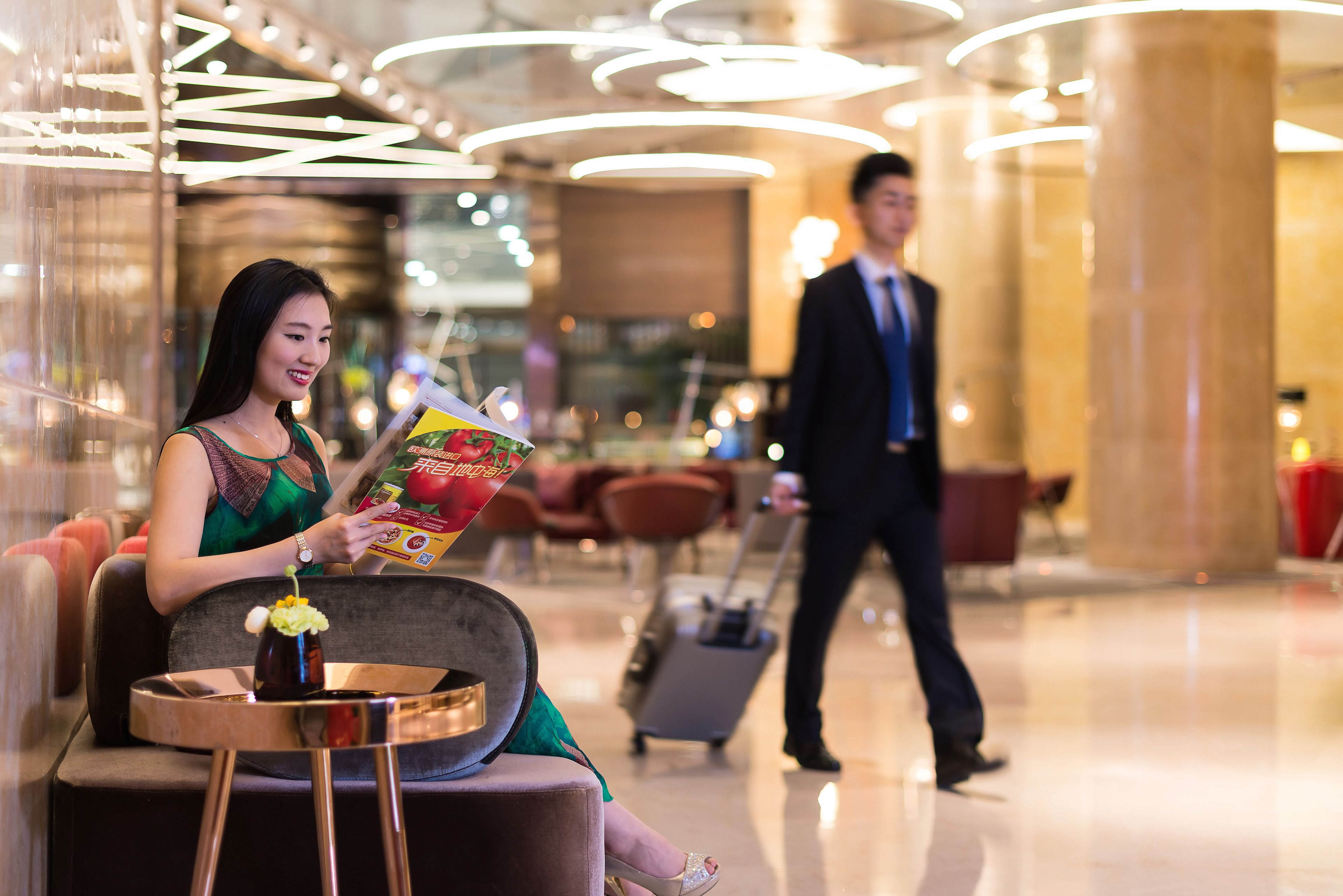 A woman sitting on a cushioned bench in a modern lobby with a vase of flowers, reading a magazine.