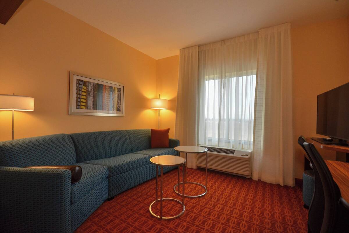 Hotellihuoneessa on sininen sohva, pieni pyöreä pöytä, televisio ja ikkuna, jossa on verhot.