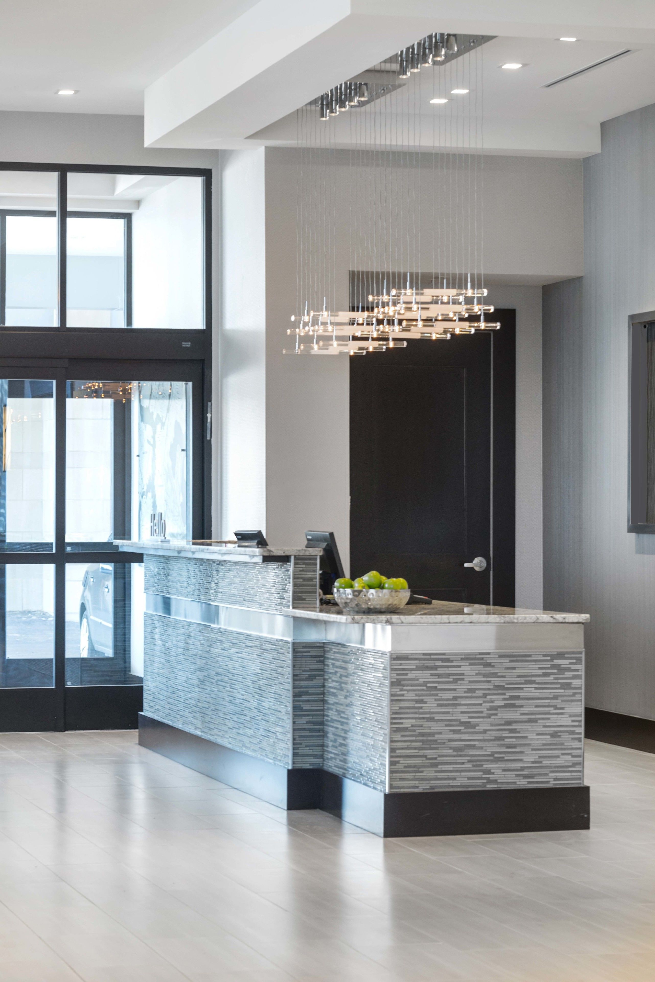 Un área de recepción moderna con entrada por puerta de cristal, un mostrador de mármol y una lámpara colgante.