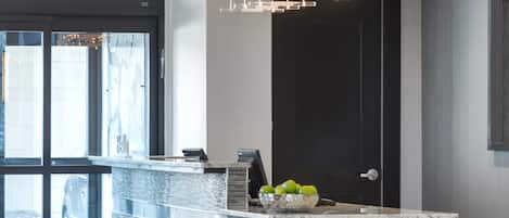 Un área de recepción moderna con entrada por puerta de cristal, un mostrador de mármol y una lámpara colgante.