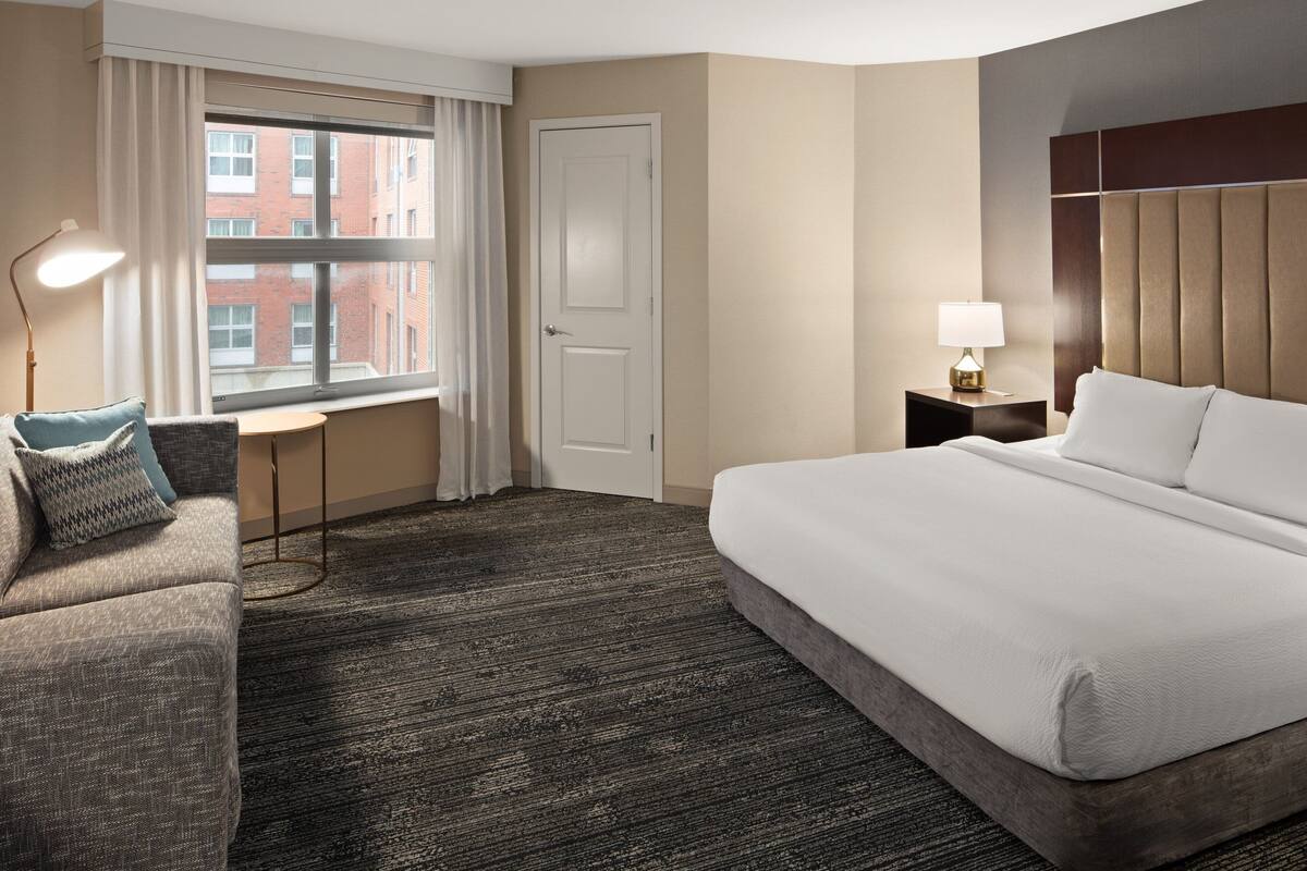 Une chambre d’hôtel avec un grand lit, un bureau, une lampe et une vue sur un immeuble par la fenêtre.