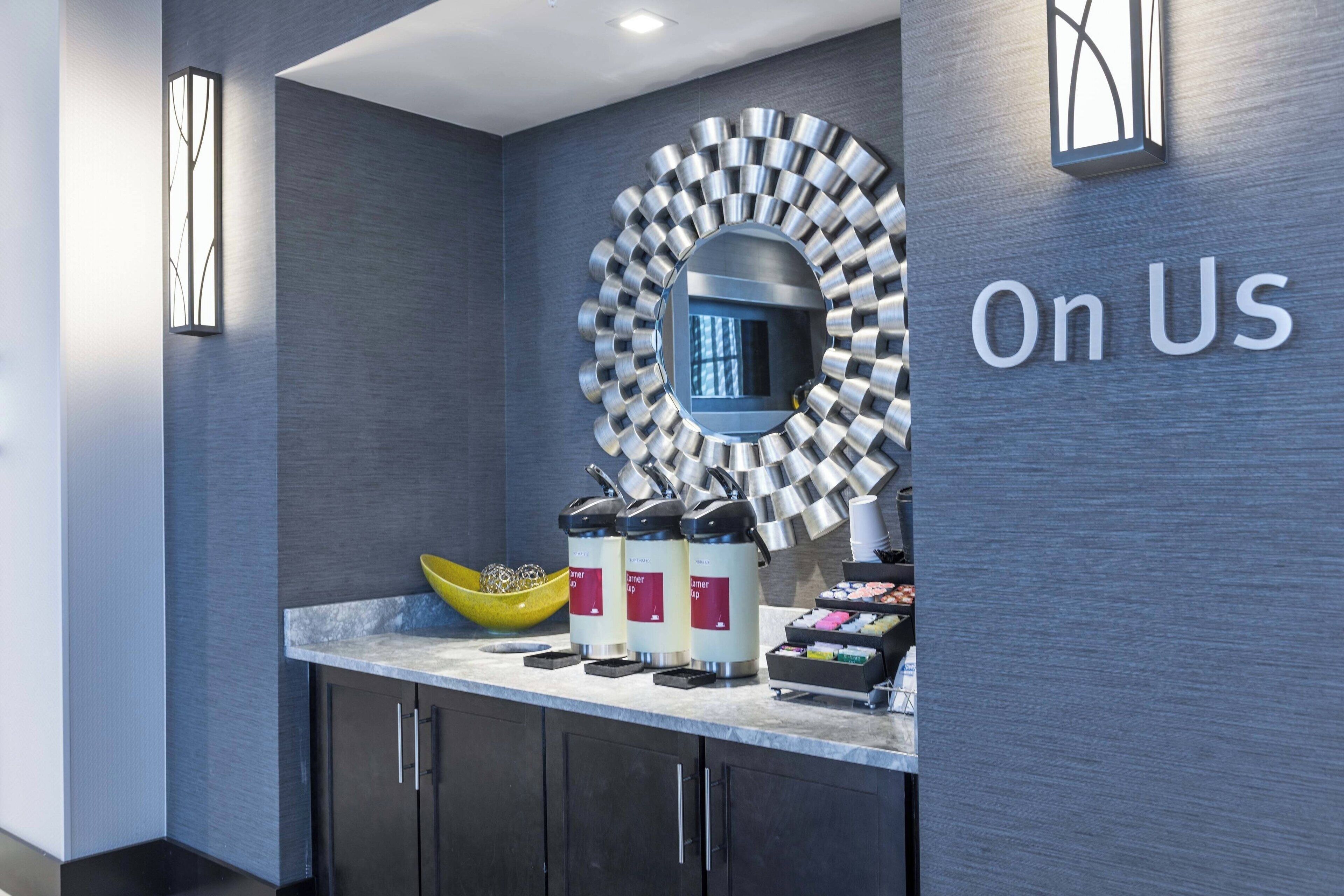 A modern hotel reception area with a coffee station, a mirror, and the text "On Us" on the wall.
