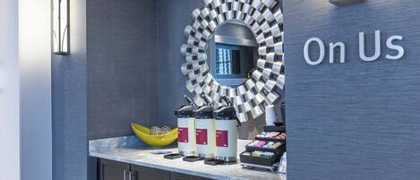 A modern hotel reception area with a coffee station, a mirror, and the text "On Us" on the wall.