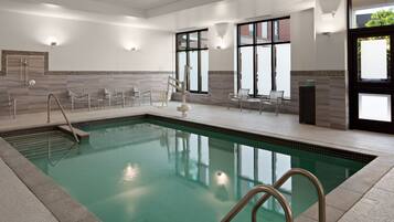A modern indoor swimming pool with clear water, surrounded by tiled walls and a seating area with chairs.