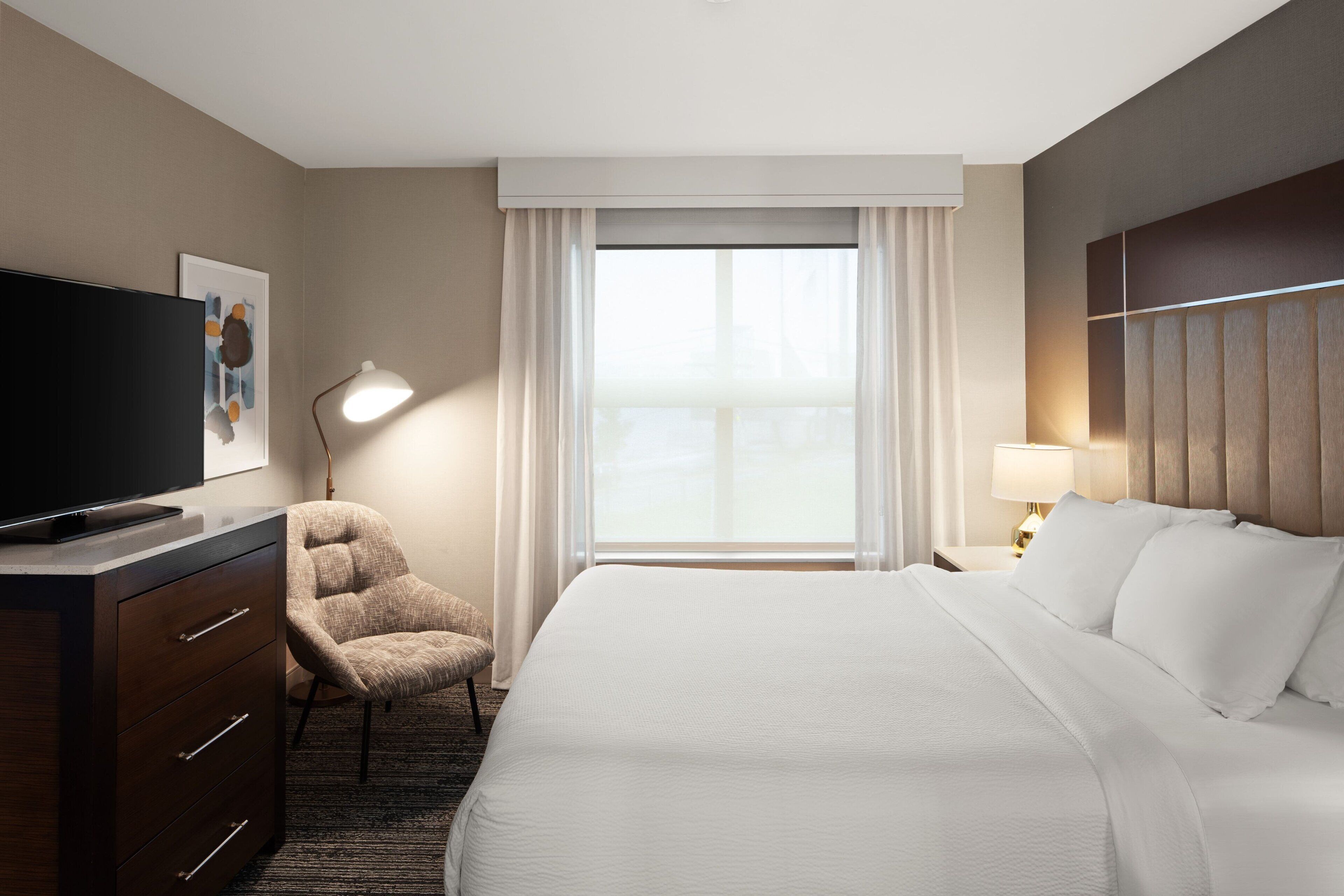 A hotel room with a bed, a TV, a desk with drawers, a chair, a lamp, and a window with curtains.