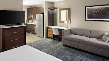 A hotel room with a bed, a TV, a kitchenette, a desk, a chair, and a sofa.