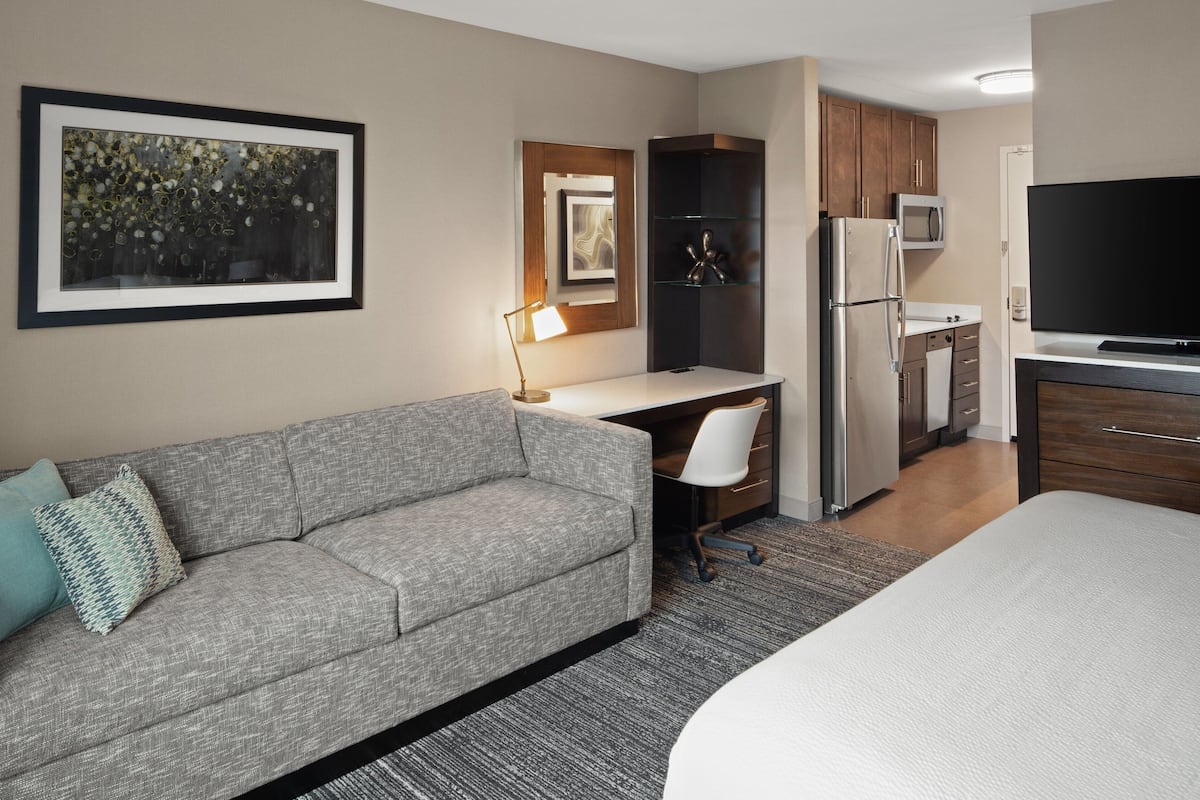 Une chambre d’hôtel moderne avec un canapé gris, un bureau avec une lampe, un lit, une télévision et une kitchenette équipée d’un réfrigérateur et d’un four à micro-ondes.