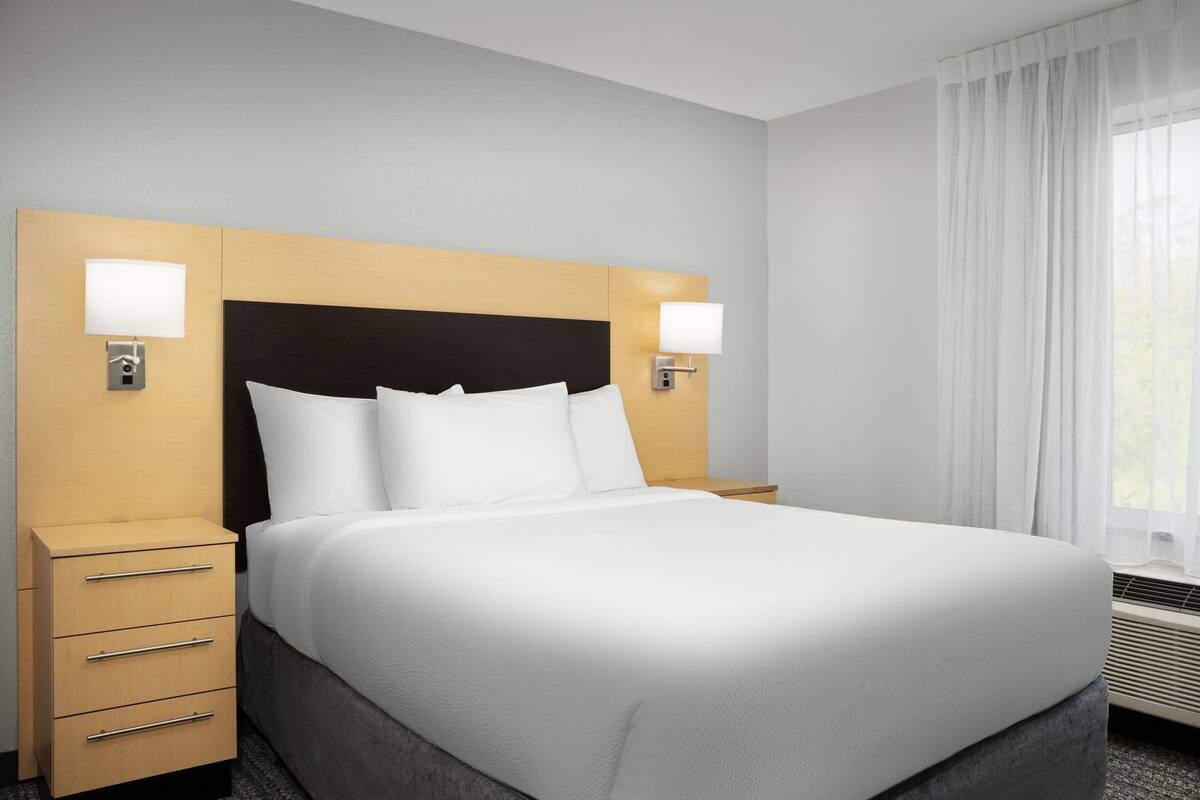 Ein Hotelzimmer mit einem großen Bett, zwei Nachttischen, einem Holz-Kopfteil und einem Fenster mit Vorhängen.