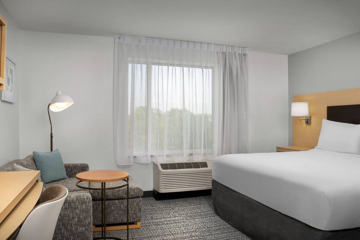 Ein Hotelzimmer mit Bett, Schreibtisch, Stuhl, einem kleinen Tisch, einer Lampe und einem Fenster mit durchsichtigen Vorhängen.