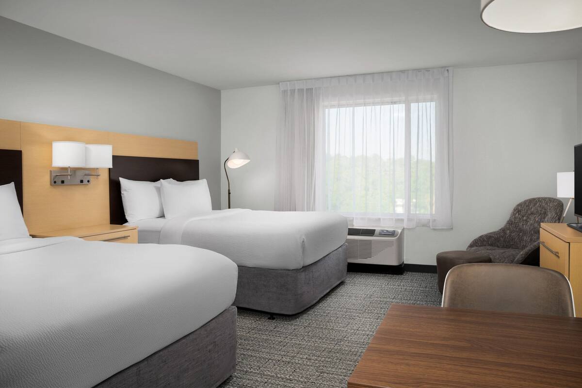 Ein Hotelzimmer mit zwei Betten, einem Schreibtisch, einem Stuhl, einer Lampe und einem fensterbelaubten Fenster.