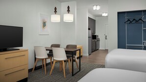 Une chambre d’hôtel moderne dotée d’une télévision à écran plat, d’un coin repas avec une table et des chaises, d’une kitchenette et d’une armoire avec espace pour suspendre les vêtements.