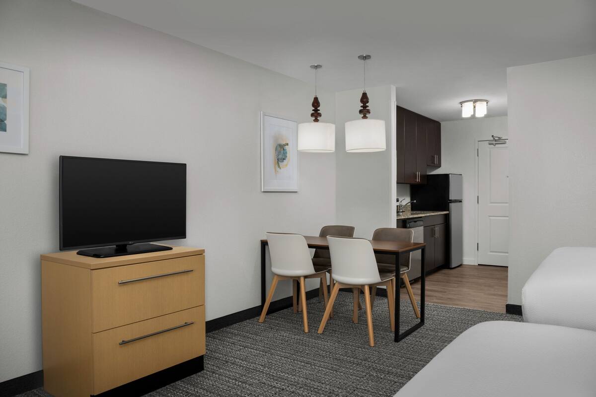 Ein modernes Hotelzimmer mit Flachbildfernseher, einem Holzschrank, einem Esstisch mit Stühlen und einer Küchenzeile.