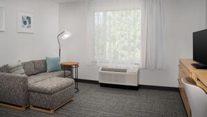Un salon moderne comprenant un canapé d’angle, un lampadaire, une grande fenêtre avec des rideaux légers, un téléviseur à écran plat et un système de climatisation intégré.