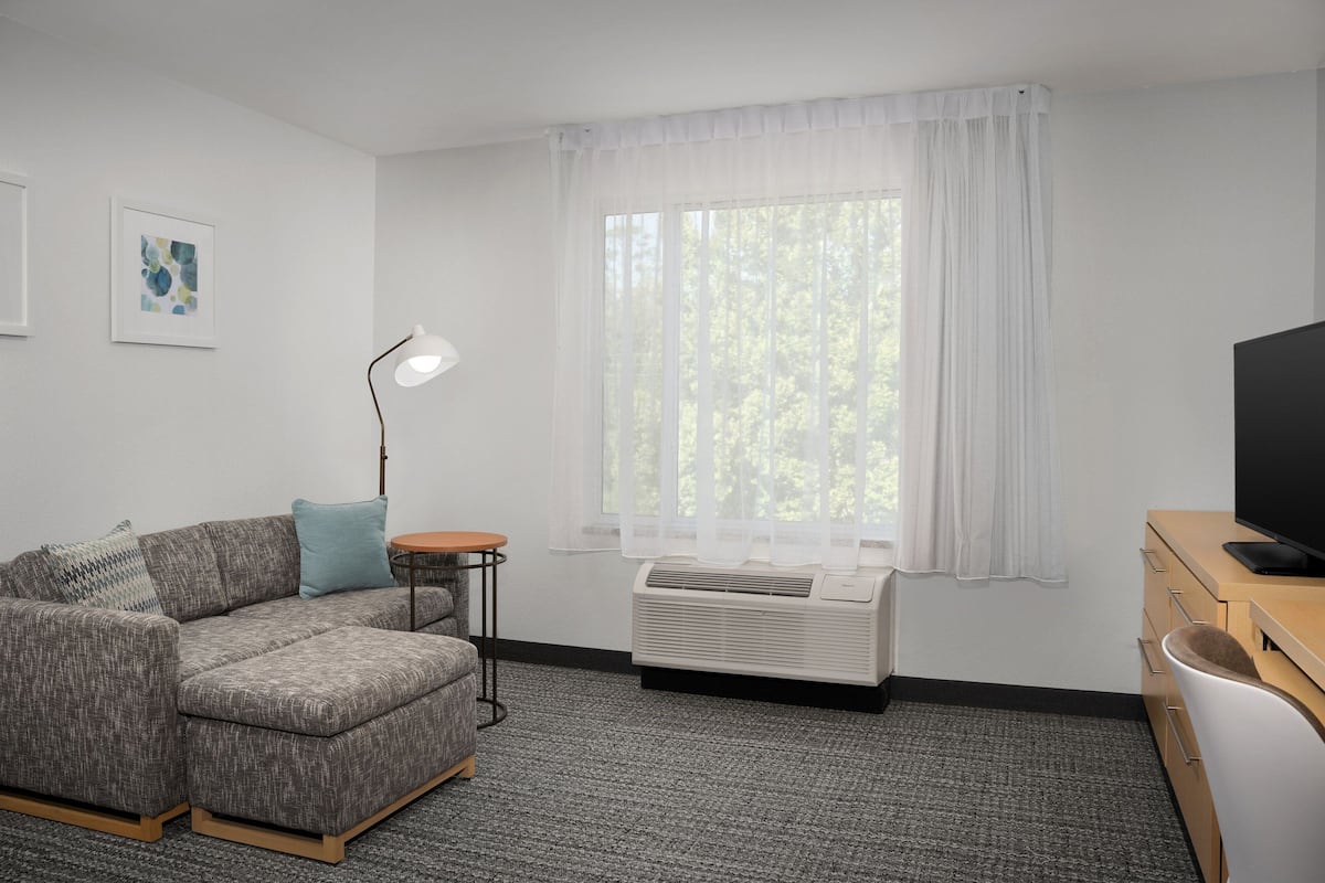 Ein modernes Wohnzimmer mit Ecksofa, Stehlampe, großem Fenster mit transparenten Vorhängen, Flachbildfernseher und integrierter Klimaanlage.