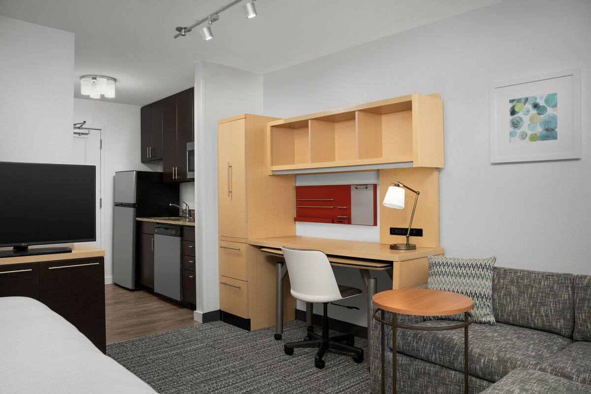 Ein kompaktes Hotelzimmer mit Küchenzeile, einem Bett, einem Schreibtisch mit Stuhl, einem kleinen Tisch und einer Couch.